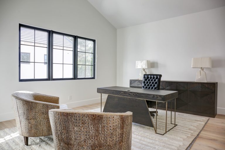 A minimalist home office design that features a sleek black desk with gold accents, a tufted leather chair, two textured armchairs, and a dark sideboard with matching table lamps.